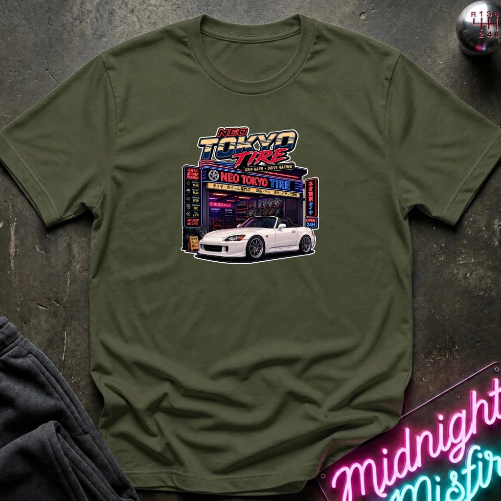 An olive green short-sleeve t-shirt with a colorful graphic print of a white sports car parked in front of a neon 'Neo Tokyo Tire' shop, laid flat on a dark textured surface.