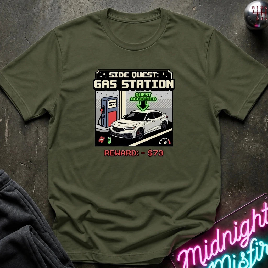 An olive green crewneck t-shirt featuring a retro video game style graphic of a gas station quest with a white car and a $73 reward, laid flat on a dark textured surface.