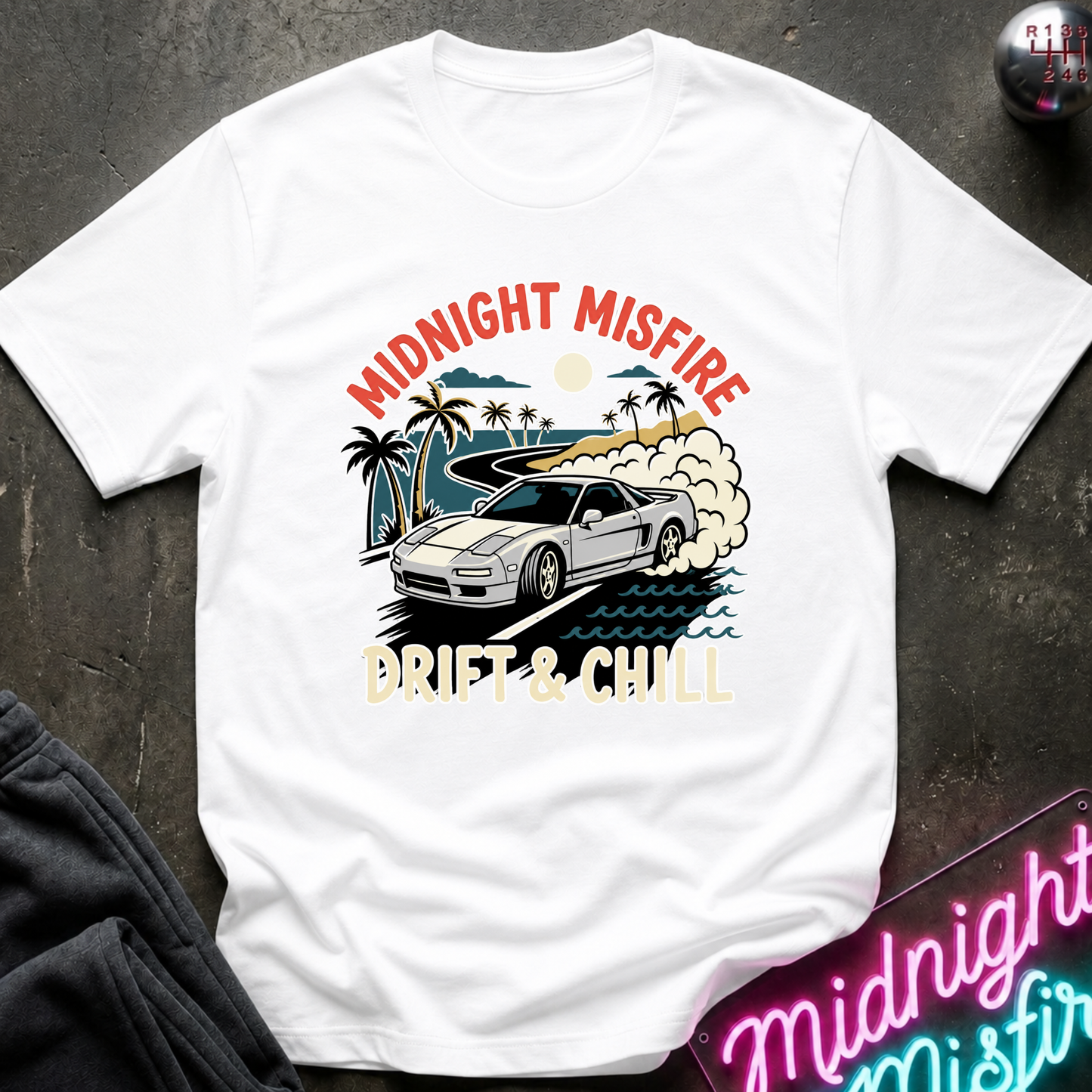 A white short-sleeve graphic T-shirt laid flat on a dark textured surface. The shirt features a colorful front print with the words 'MIDNIGHT MISFIRE' and 'DRIFT & CHILL' around an illustration of a silver sports car on a beach with palm trees and waves.