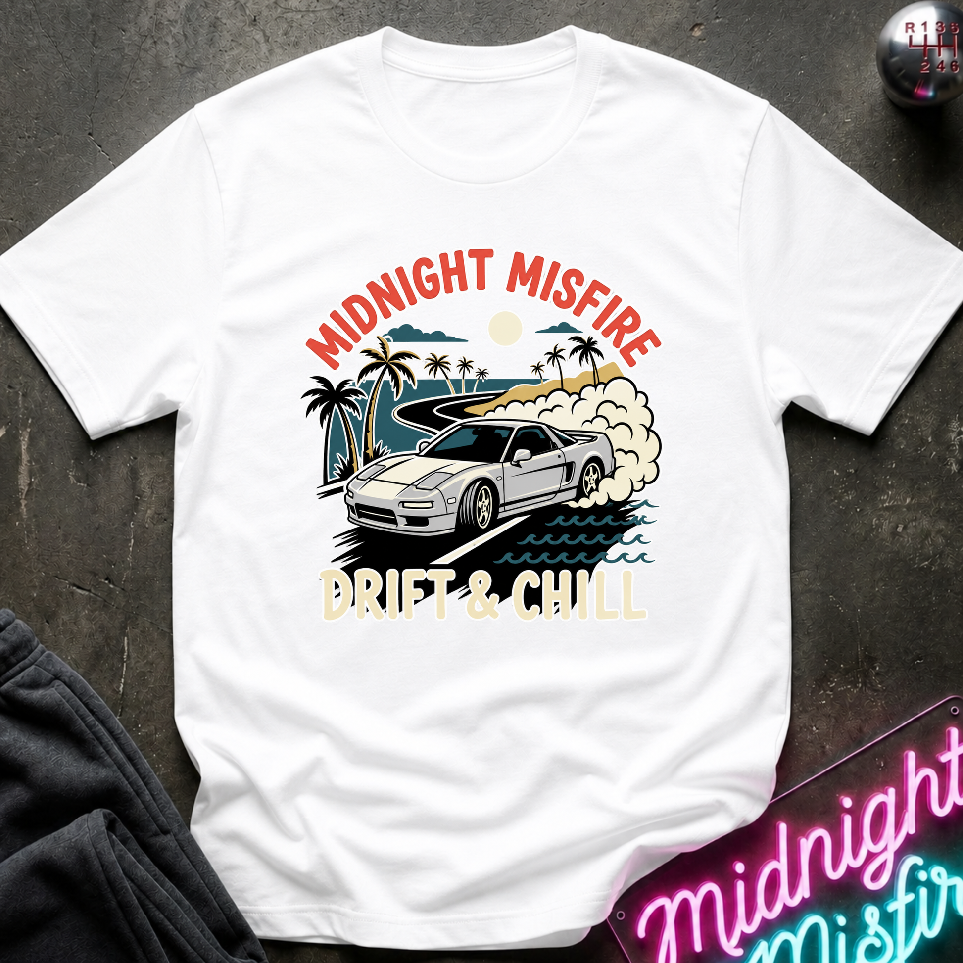 A white short-sleeve graphic T-shirt laid flat on a dark textured surface. The shirt features a colorful front print with the words 'MIDNIGHT MISFIRE' and 'DRIFT & CHILL' around an illustration of a silver sports car on a beach with palm trees and waves.