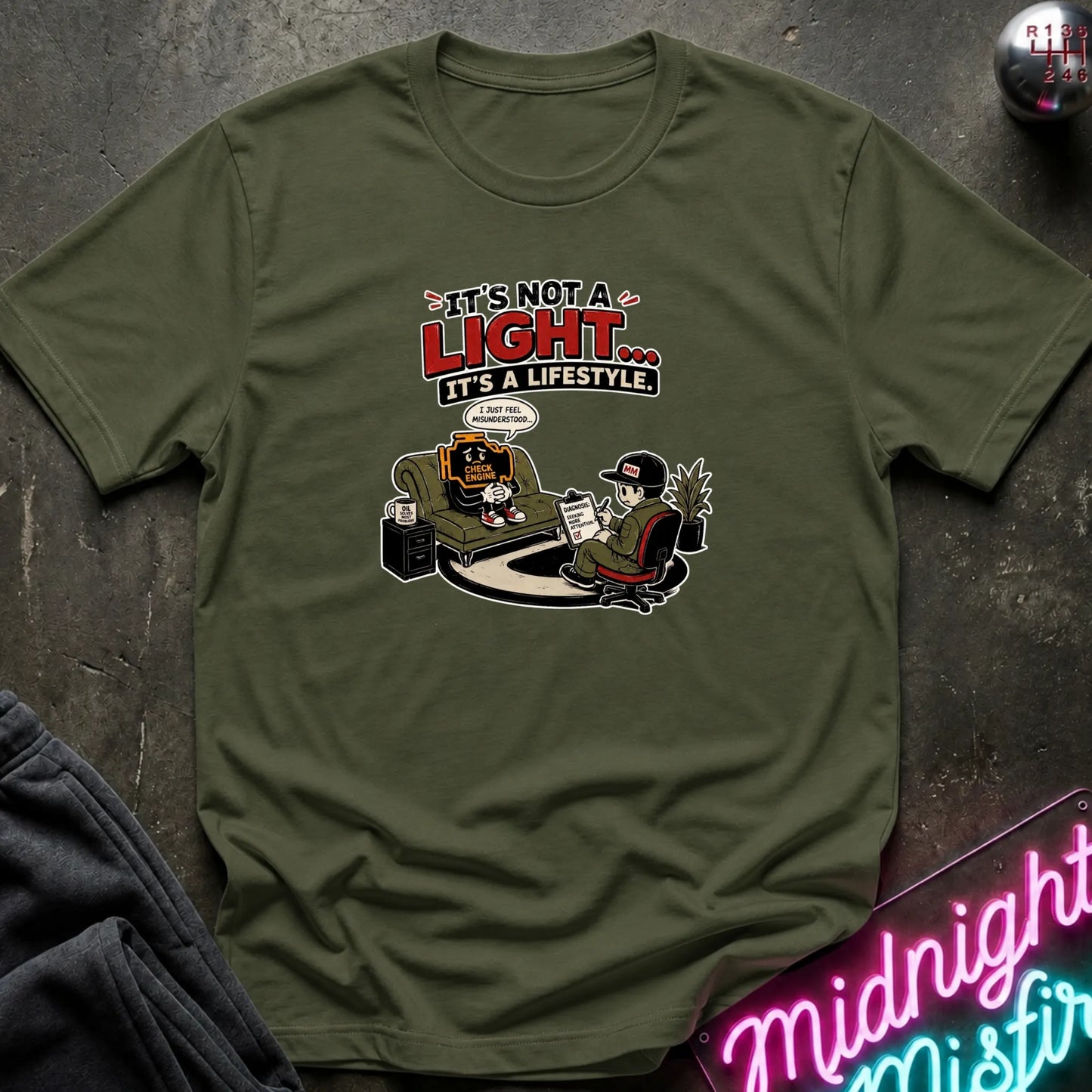 An olive green graphic t-shirt laid flat on a dark textured surface. The graphic features a cartoon illustration of a person sitting in a chair with a mug, with text above reading 'IT'S NOT A LIGHT.