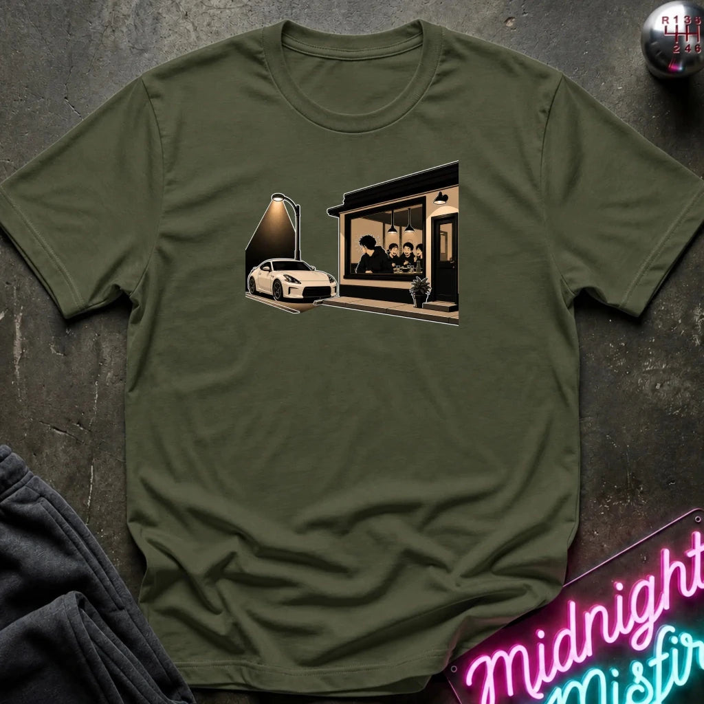 An olive green crewneck t-shirt laid flat on a dark textured surface, featuring a graphic print of a white sports car parked outside a dimly lit cafe. 