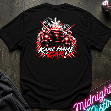 A black short-sleeve t-shirt laid flat on a dark textured surface, featuring a large back graphic of a black sports car with red headlights, smoke effects, and the text 'KAME HAME CAR!' in red and white. A neon sign and folded dark fabric are partially visible around it.