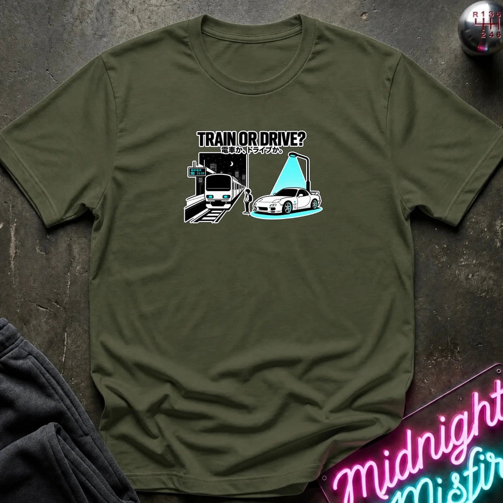 olive graphic t-shirt laid flat on a dark textured surface. The graphic features a train and a car with a person.