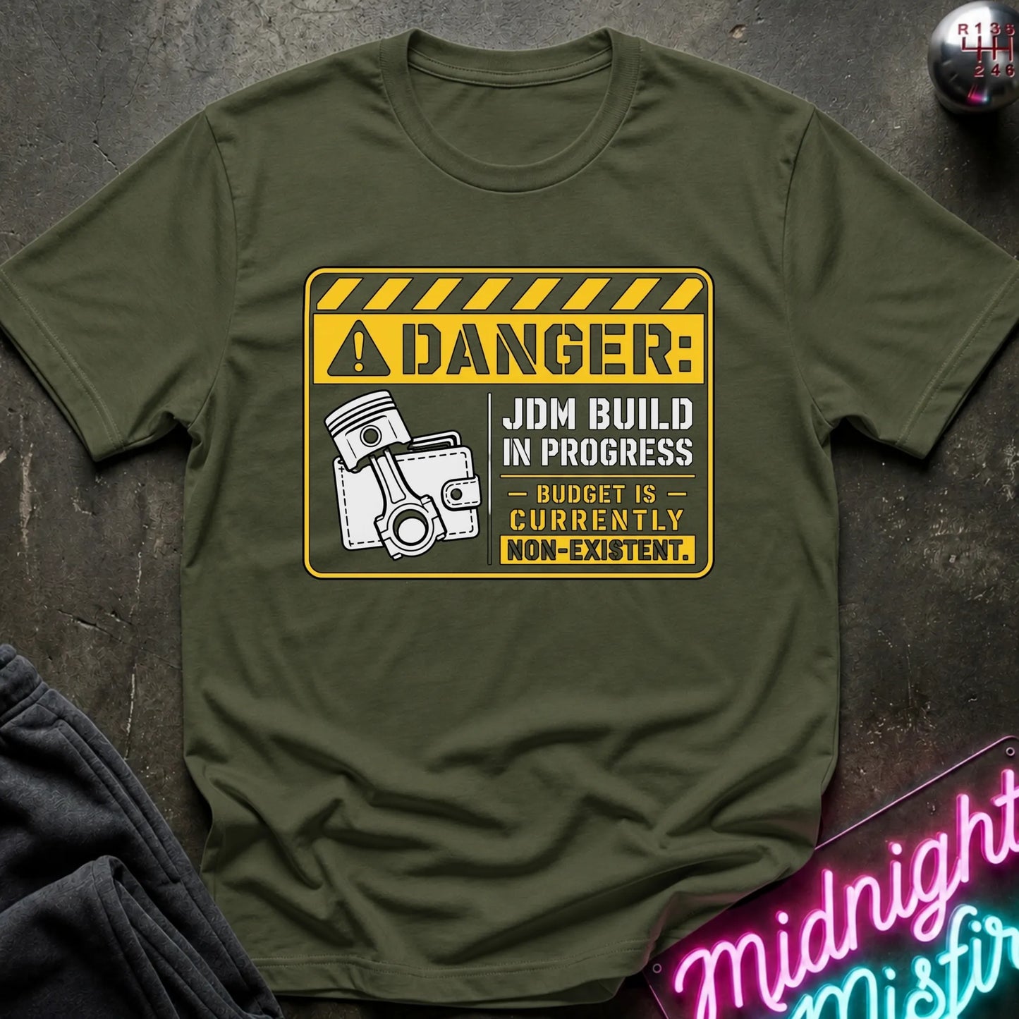 An olive green  short-sleeve graphic T-shirt laid flat on a dark textured surface. The shirt features a yellow and white warning-sign style print with the text 'DANGER: JDM BUILD IN PROGRESS - BUDGET IS - CURRENTLY NON-EXISTENT'.