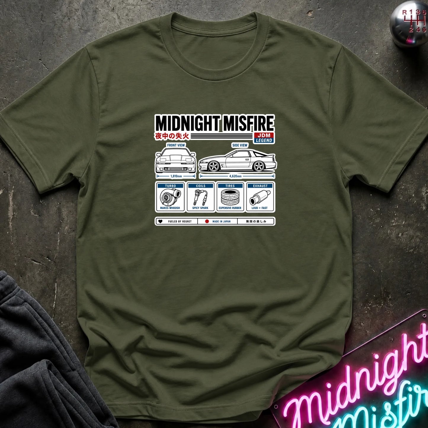 An olive green short-sleeve t-shirt with a white and black graphic print titled 'MIDNIGHT/MISFIRE' featuring a car diagram and Japanese text, laid flat on a dark textured surface with a neon sign and dark fabric partially visible.