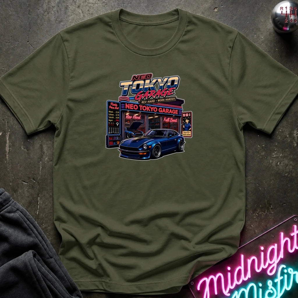 An olive green crewneck t-shirt featuring a colorful graphic print of a blue sports car in front of a 'Neo Tokyo Garage' sign, styled flat on a dark textured surface with a neon sign and dark pants partially visible.