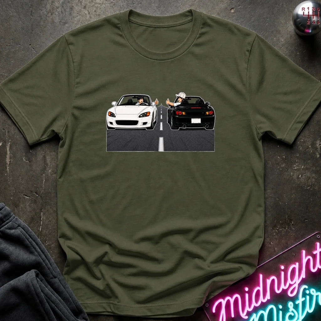 An olive green crewneck t-shirt featuring a graphic print of a white convertible and a black sports car on a road, styled flat on a dark textured background with a neon sign and sweatpants.