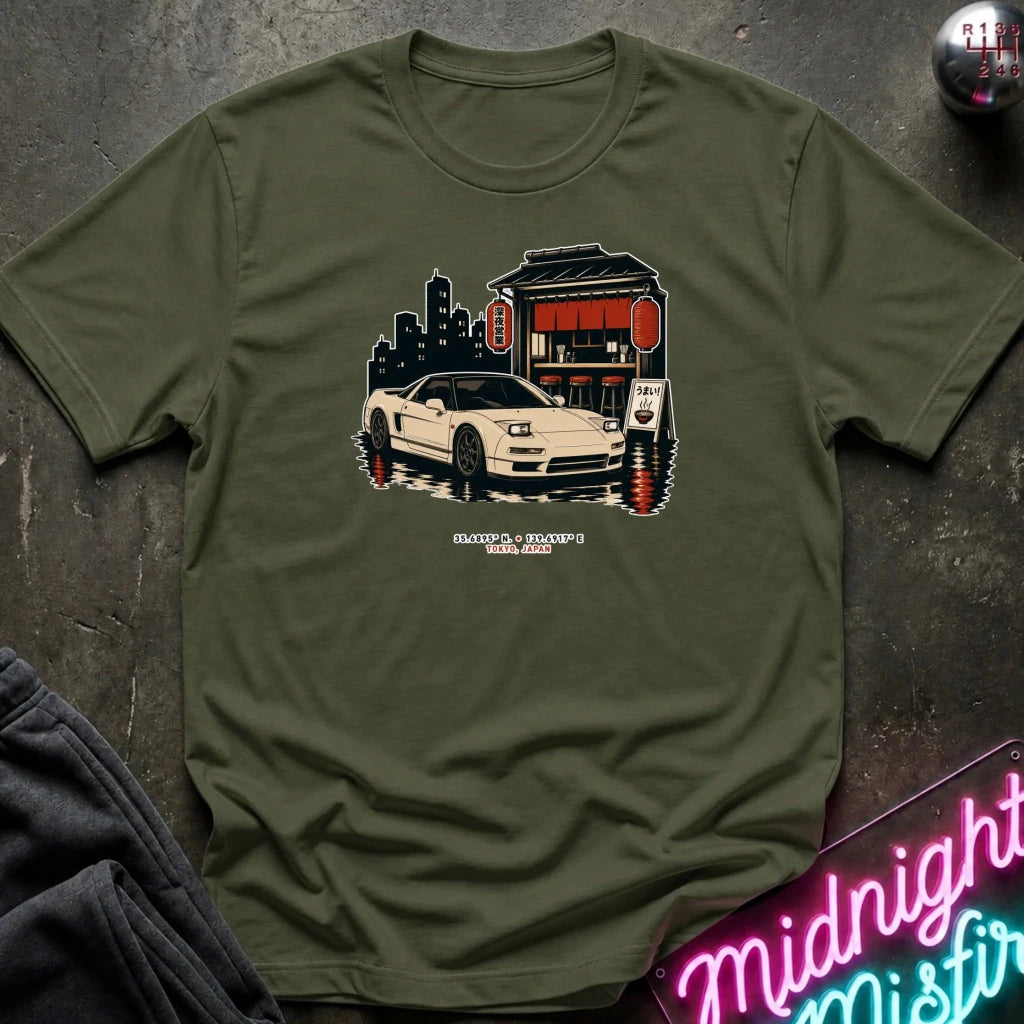 An olive green graphic t-shirt laid flat on a dark textured surface, featuring a retro illustration of a white sports car parked in front of a convenience store with a neon sign. 