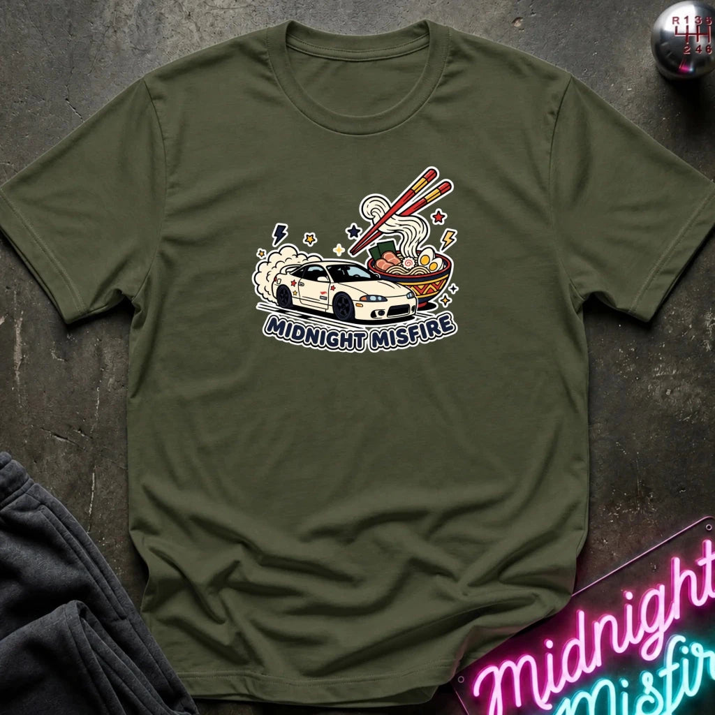 An olive green crewneck t-shirt laid flat on a dark textured surface, featuring a graphic of a white sports car driving into a ramen bowl with chopsticks, and the text 'MIDNIGHT MISFIRE'.