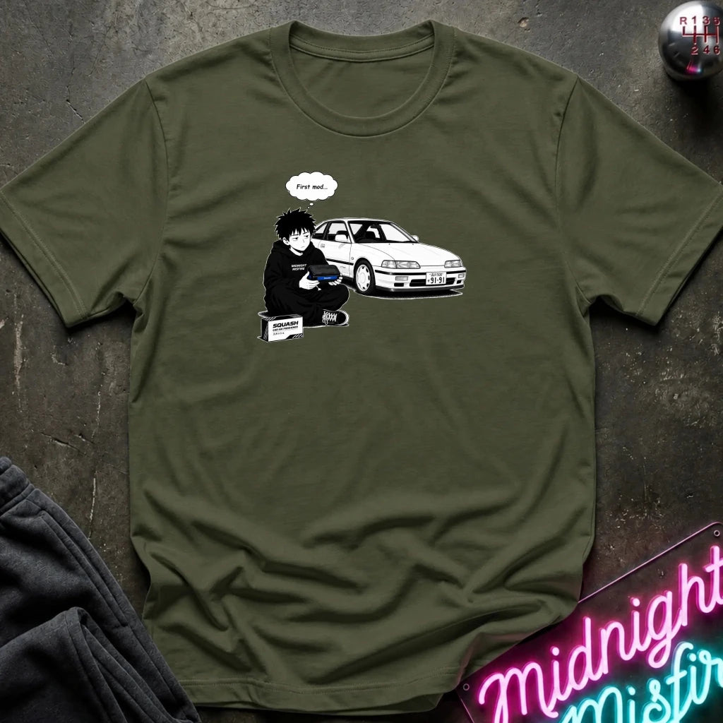 An olive green crewneck t-shirt laid flat on a dark textured surface, featuring a graphic print of a person sitting next to a white car with a speech bubble 