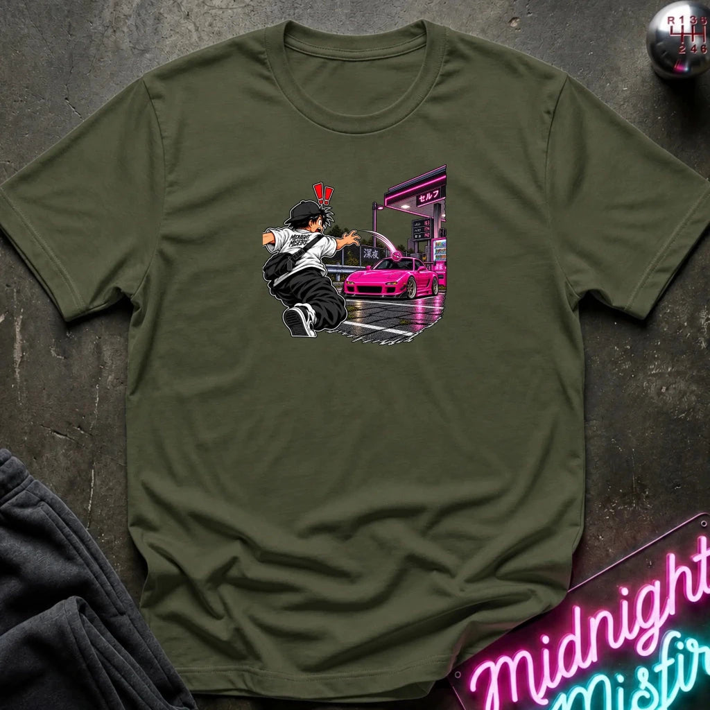 An olive green short-sleeve crewneck t-shirt laid flat on a dark textured surface. The center of the shirt features a graphic print of an anime character in a black and white outfit with a hat, next to a sports car 
