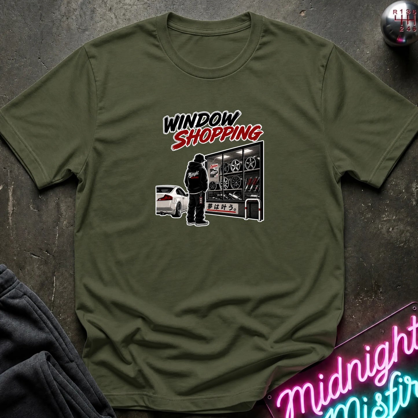 An ollve green short-sleeved t-shirt  that has a cars parked and a person checking out windows of parts.