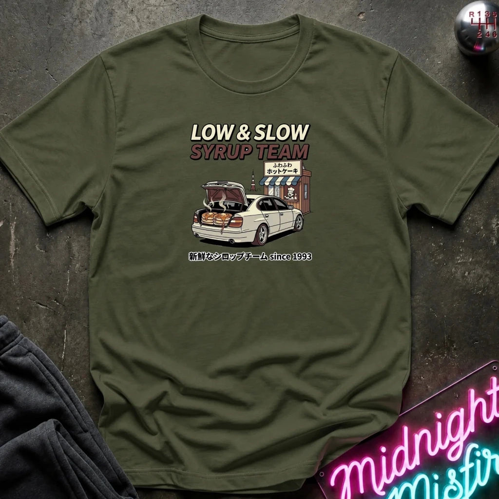 An olive green crewneck t-shirt with a graphic print of a white car and the text 'LOW & SLOW SYRUP TEAM' in white and red, laid flat on a dark textured surface with a neon sign and dark pants partially visible.