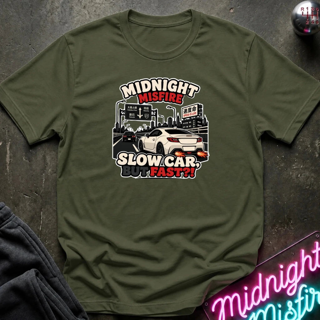 An olive green short-sleeve t-shirt with a graphic print of a white sports car and the text 'MIDNIGHT MISFIRE SLOW CAR, FAST!!' in red, white, and black, laid flat on a dark textured surface.