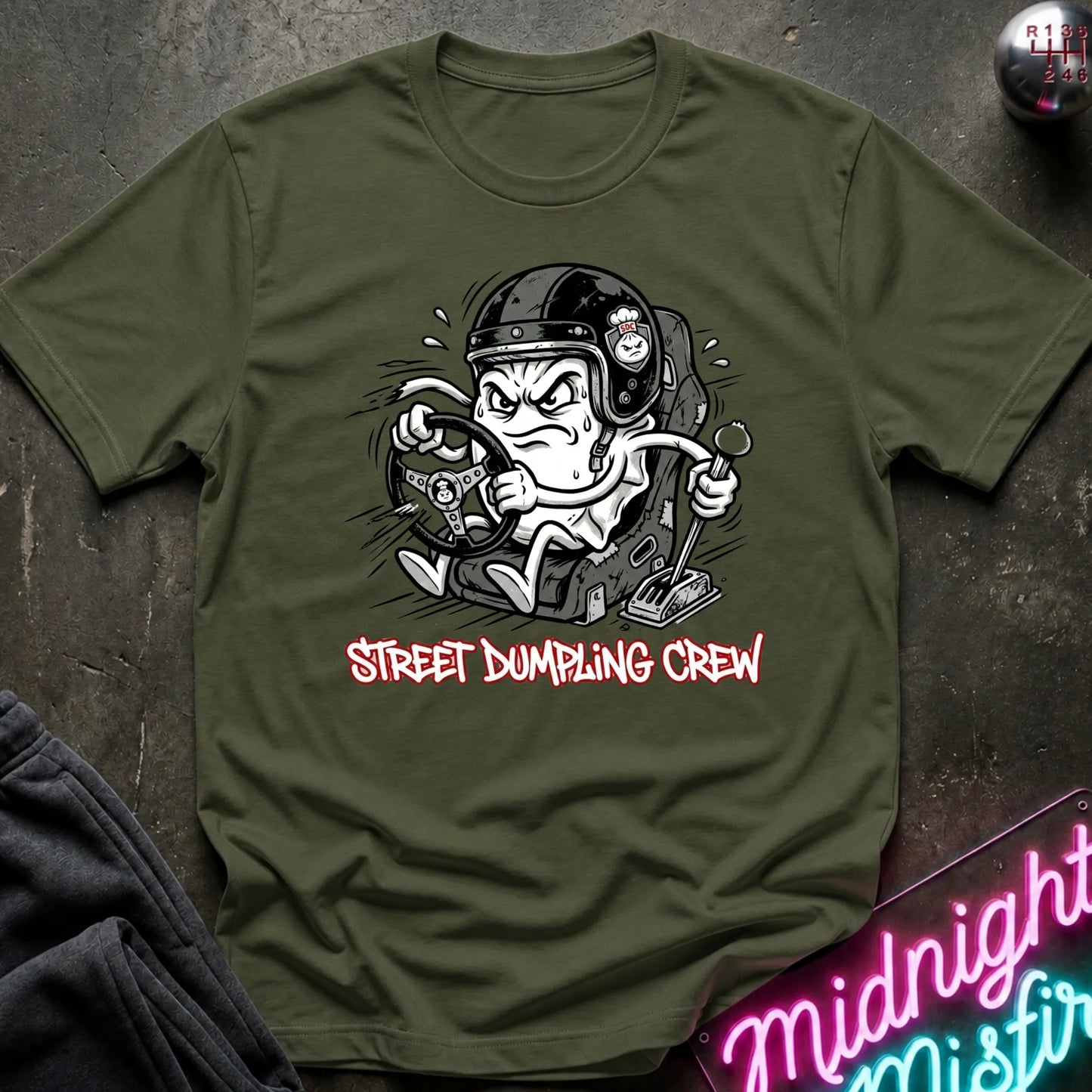 An olive green graphic t-shirt laid flat on a dark textured surface. The shirt features a cartoon illustration of a street-dumping character in a helmet with the text 'STREET DUMPING CREW' printed below in red and white.