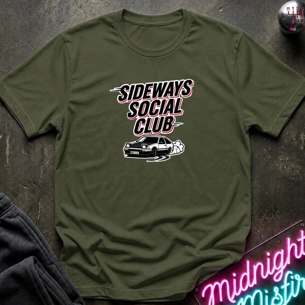 An olive green crewneck t-shirt with a graphic print reading 'SIDEWAYS SOCIAL CLUB' above a drifting car and smoke, laid flat on a dark textured surface with a neon sign and dark pants partially visible.