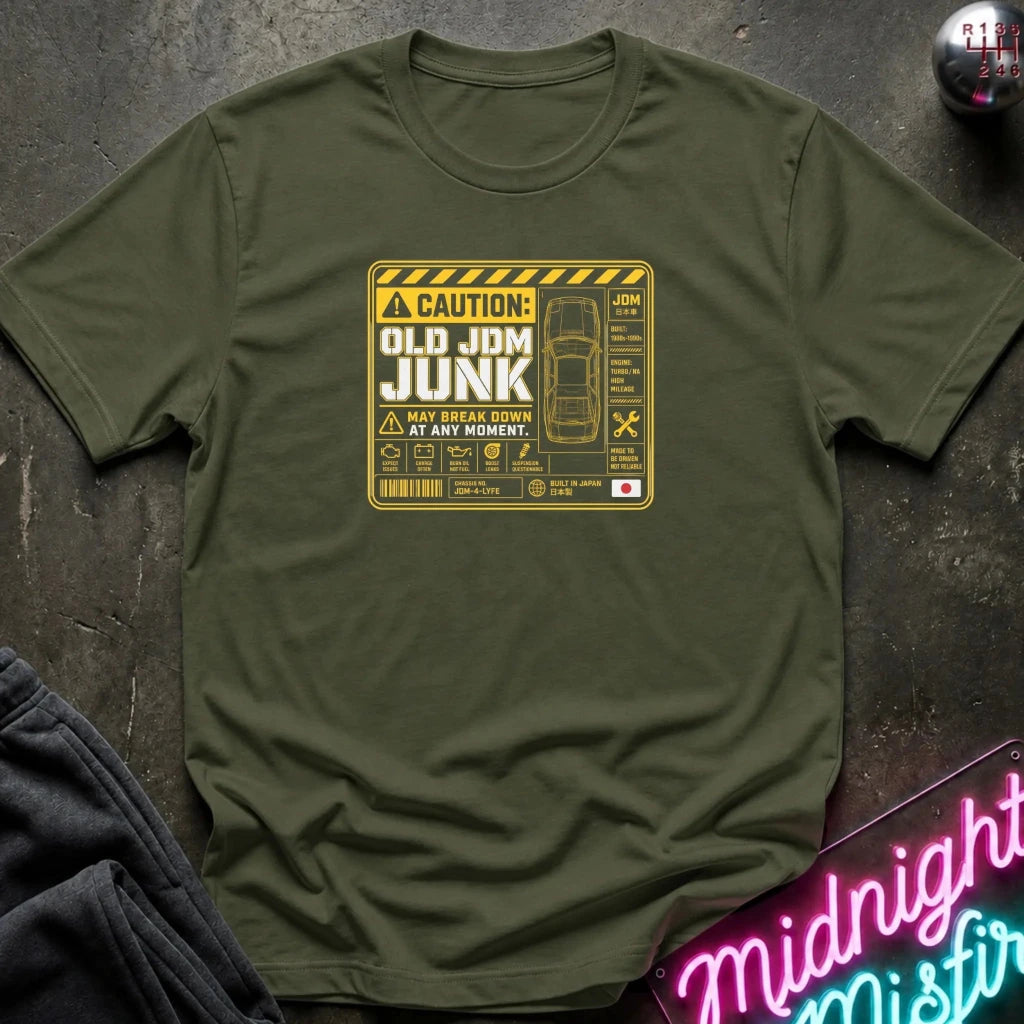 An olive green crewneck t-shirt with a yellow and black caution-style graphic that reads 'OLD JDM JUNK' and shows a car schematic, laid flat on a dark textured surface with a neon sign and dark fabric partially visible.