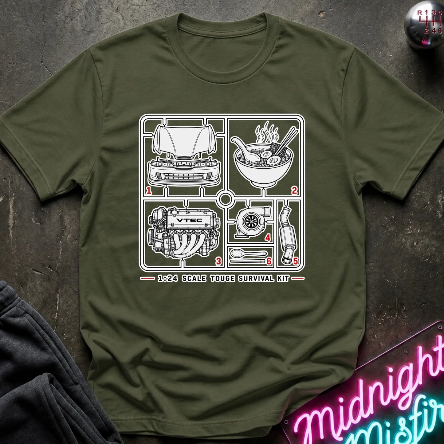 An olive green short-sleeve graphic T-shirt laid flat on a dark textured surface. The shirt features a black line-art schematic of a survival kit with numbered illustrations and the text '1:24 SCALE TOUGHE SURVIVAL KIT' printed on the chest.