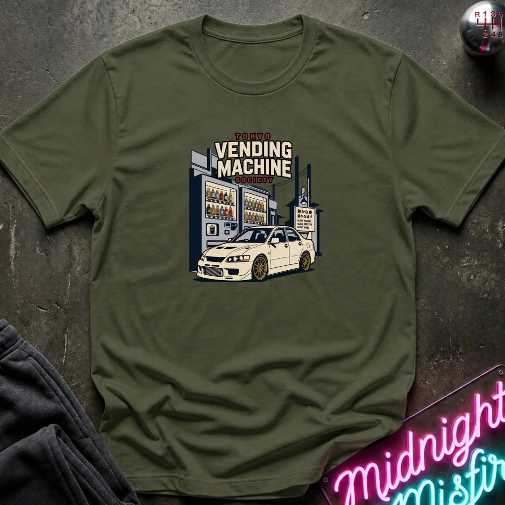 An olive green crewneck t-shirt laid flat on a dark textured surface, featuring a graphic print of a white sports car parked in front of a vending machine. The shirt is styled with a dark gray sweatshirt in the corner and a pink neon sign in the bottom right.
