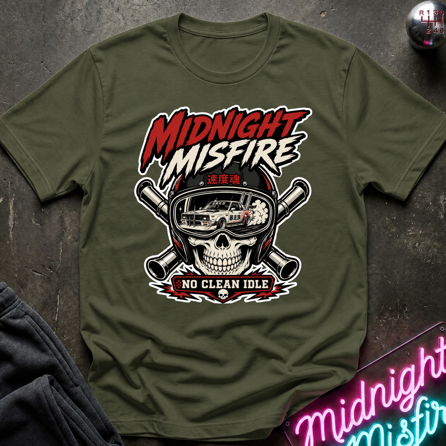 An olive green short-sleeve t-shirt with a 'Midnight Misfire' graphic on the chest, featuring a skull in a helmet, crossed pipes, and a car, displayed flat on a dark textured surface.