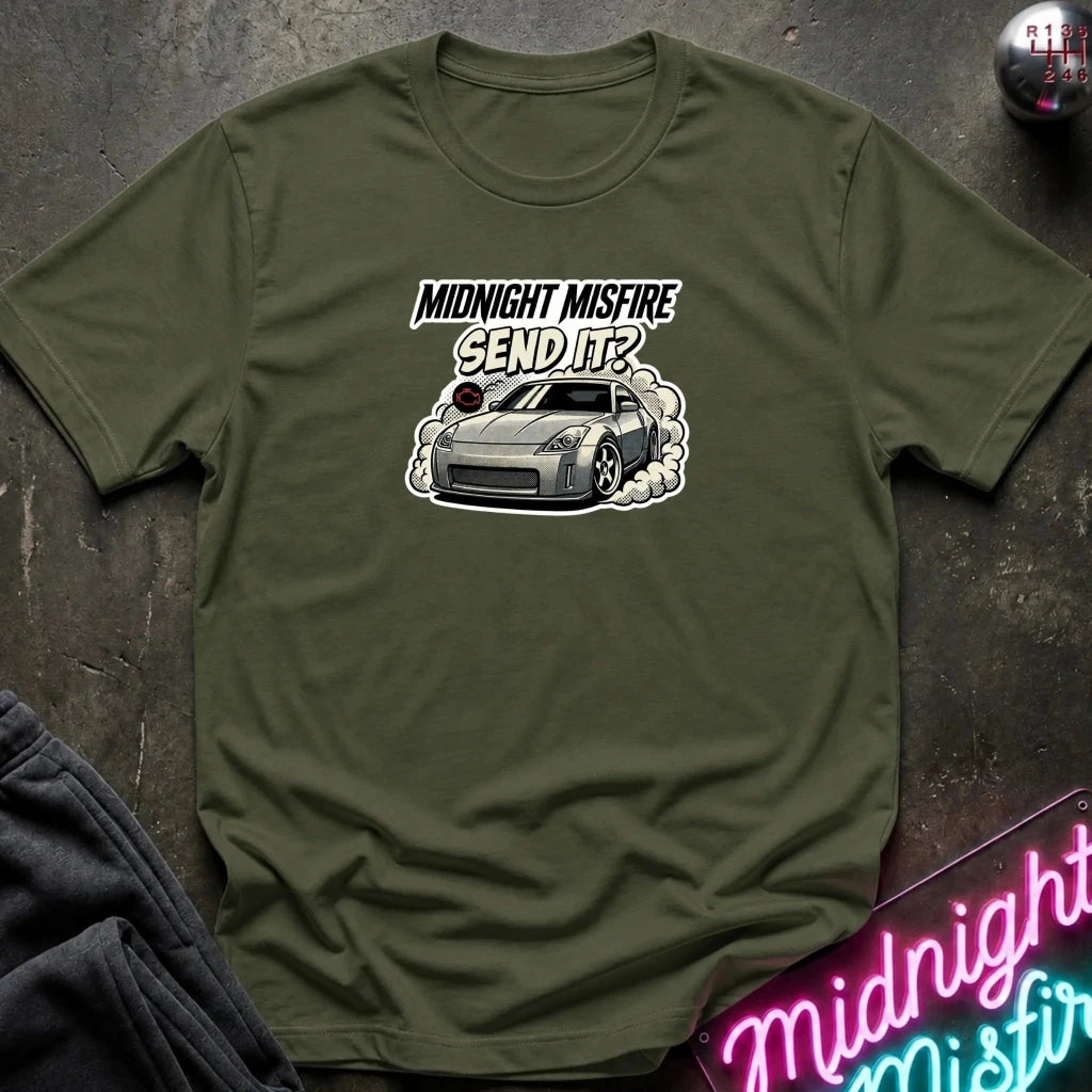 An olive green short-sleeve t-shirt with a graphic print of a silver sports car and the text 'MIDNIGHT MISFIRE SEND IT?' on the chest, laid flat on a dark textured surface with a neon sign and dark pants partially visible.