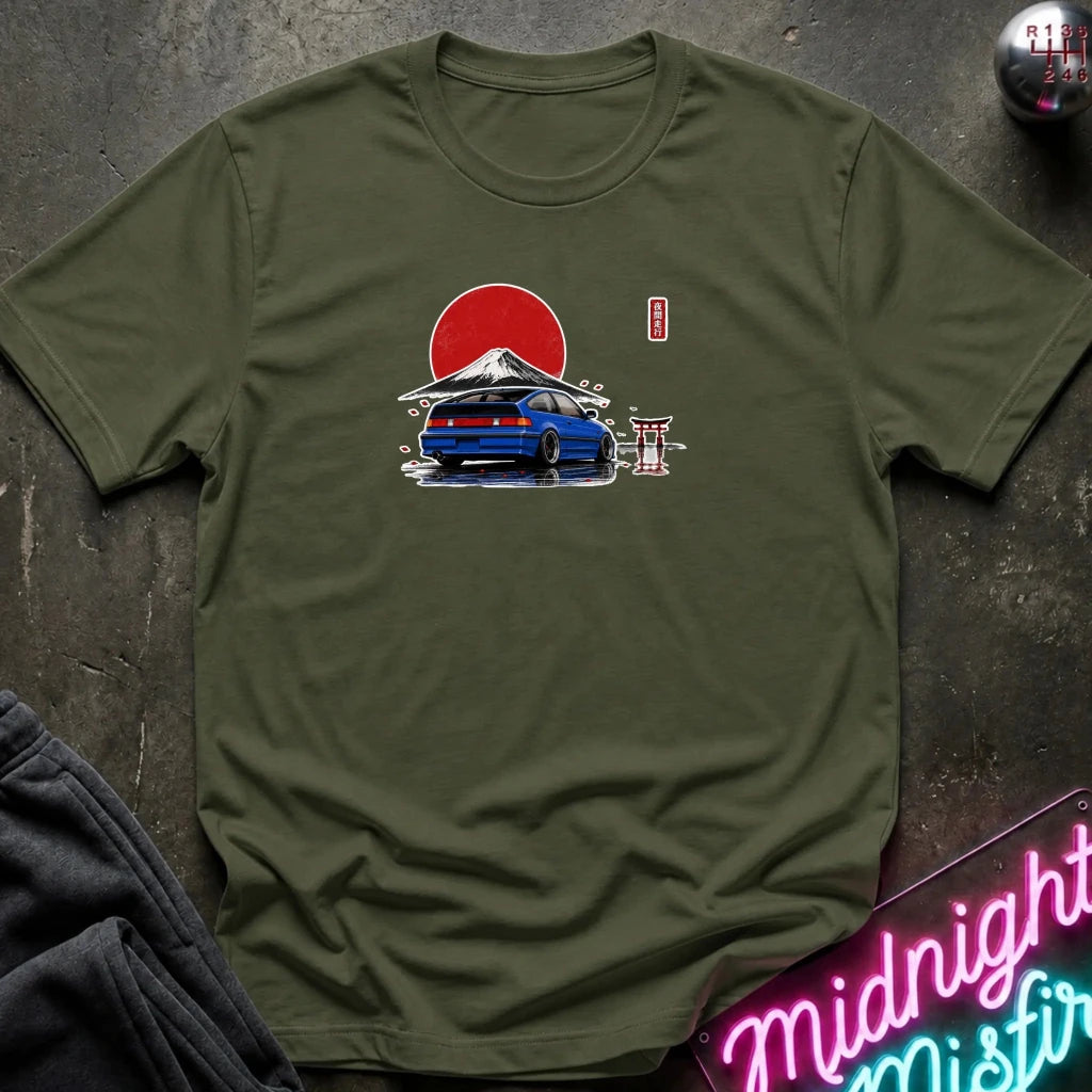 An olive green short-sleeve t-shirt laid flat on a dark textured surface, featuring a graphic print of a blue car in front of a red sun and mountain with a small red vertical flag. A neon sign and folded dark fabric are partially visible around the edges.