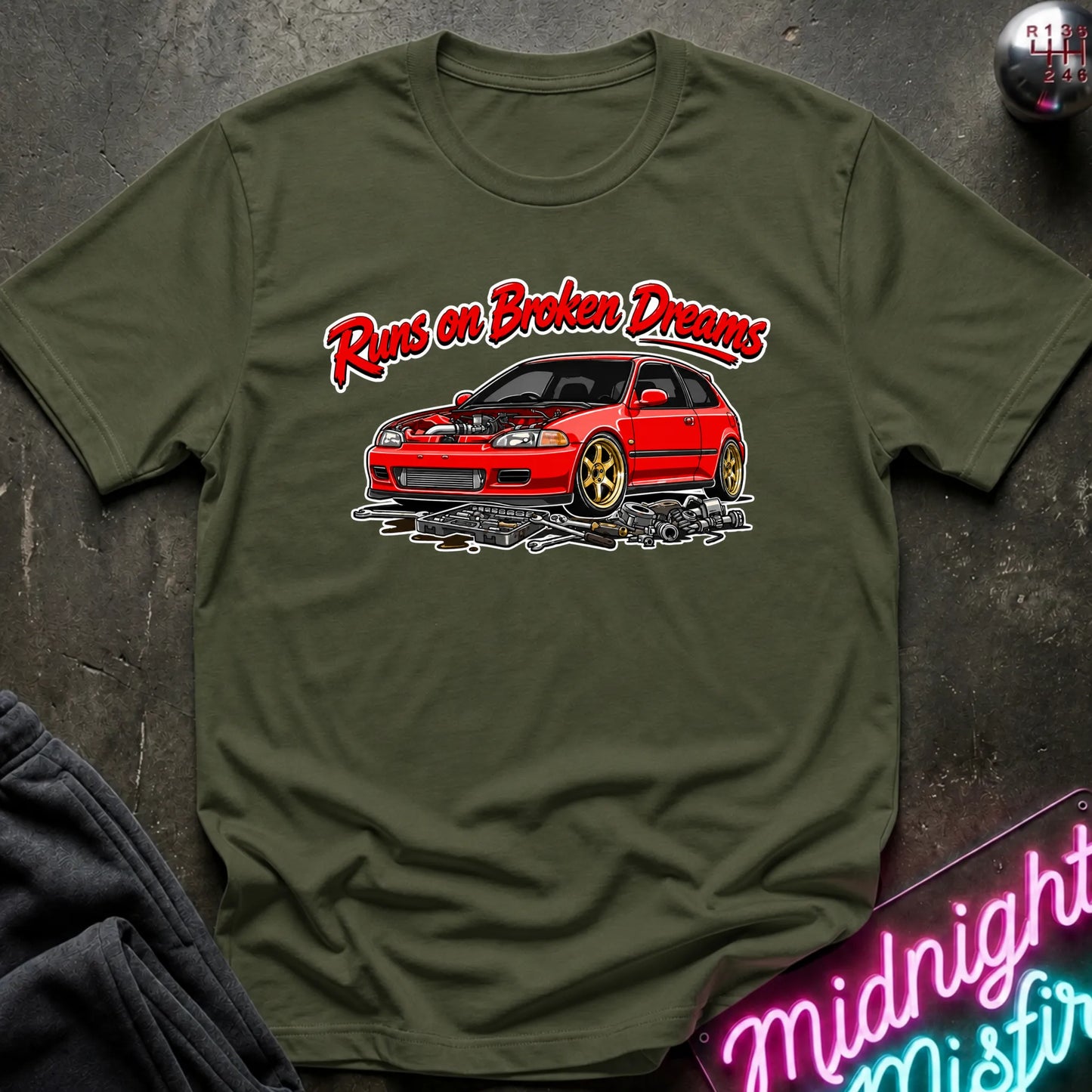 olive green graphic t-shirt laid flat on a dark textured surface. The shirt features a red sports car with gold wheels and the text 'Runs on Broken Dreams' in red and white.