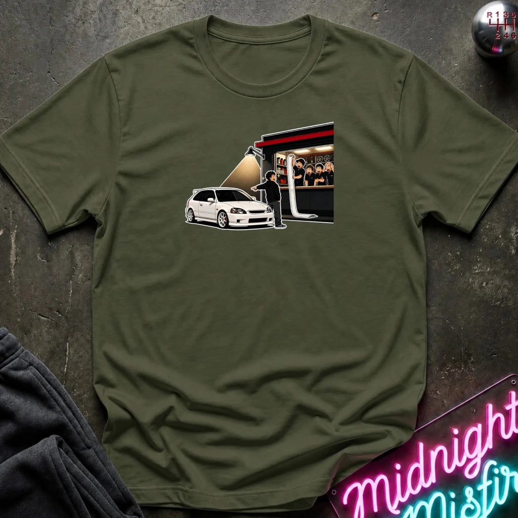 An olive green crewneck t-shirt laid flat on a dark textured surface, featuring a graphic print of a white car in front of a red and black trailer with people visible inside.