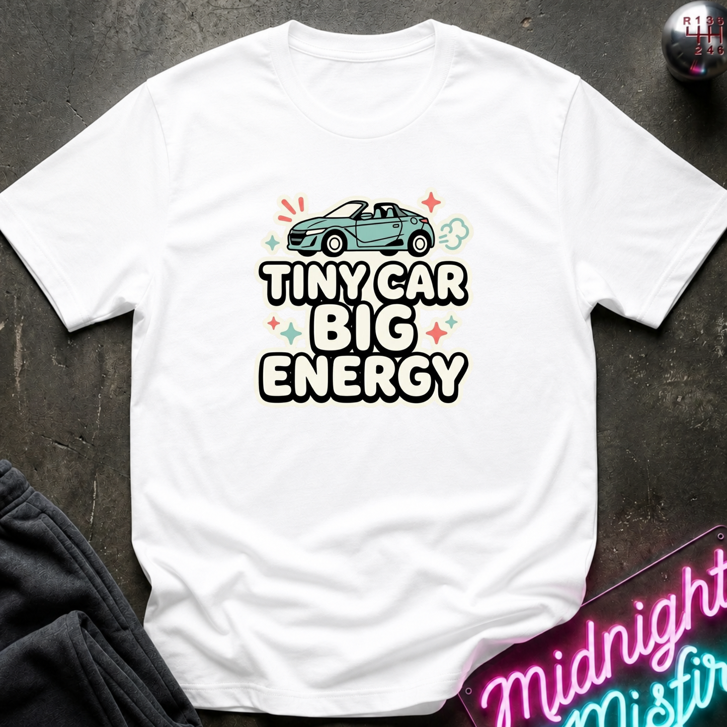 A white short-sleeve graphic T-shirt laid flat on a dark textured surface. The shirt features a light blue convertible car illustration above the text 'TINY CAR BIG ENERGY' in bold black letters with white outlines, accented by small colorful star and cloud shapes.