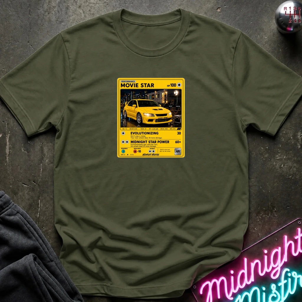 olive short-sleeve t-shirt laid flat on a dark textured surface, featuring a yellow and black retro-style graphic on the chest with a yellow car photo and text including 'MOVIE STAR'. 