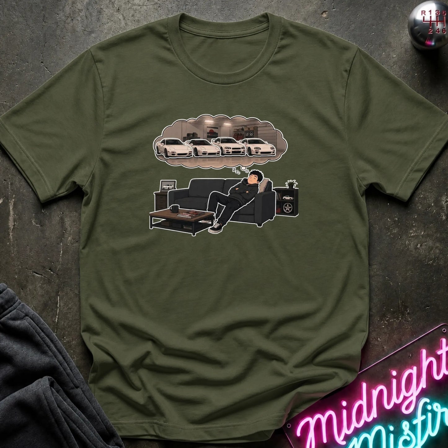 An olive green short-sleeve t-shirt laid flat on a dark textured surface, featuring a graphic print of a man lounging on a couch beneath a thought bubble showing  white cars. A neon is partially visible in the bottom right corner.