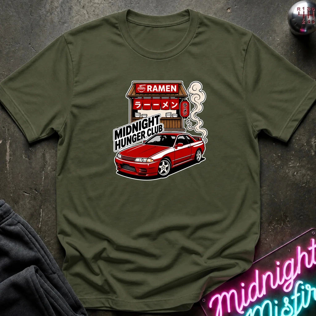 An olive green crewneck t-shirt featuring a graphic of a red sports car parked in front of a ramen restaurant with the text 'MIDNIGHT HUNGER CLUB'. The shirt is displayed flat on a dark textured surface with a neon sign and dark fabric in the corners.