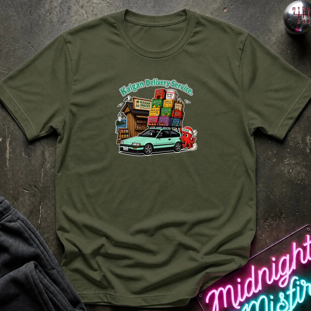 An olive green graphic t-shirt laid flat on a dark textured surface. The graphic features a light green car, and a small building with colorful signs under the text 'Kaigan Delivery Service'.