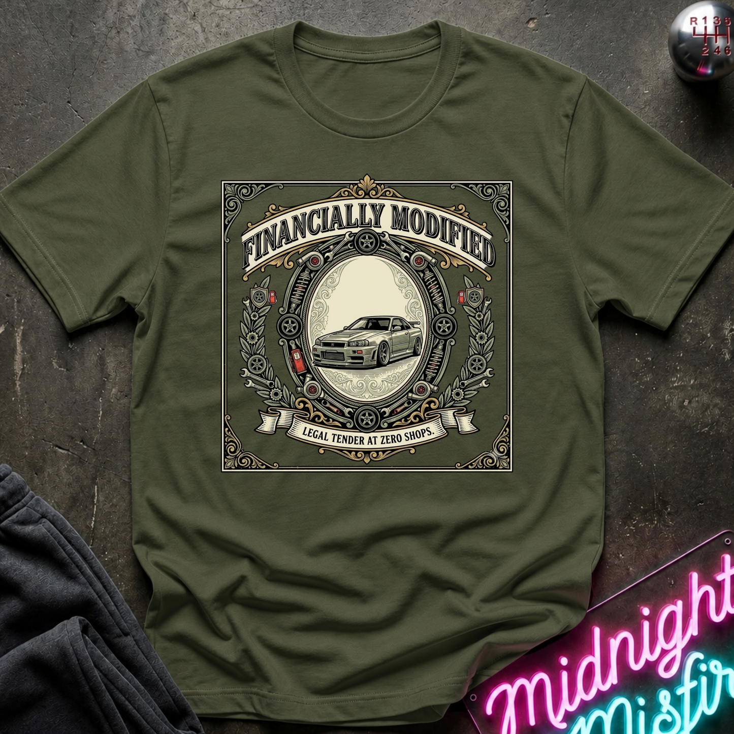 An olive green short-sleeve t-shirt with a vintage-style graphic print that reads 'FINANCALLY MODIFIED' and 'LOCAL TENDER AT ZERO SHOPS' around an illustration of a classic car. The shirt is laid flat on a dark textured surface with a neon sign and dark fabric partially visible in the corners.