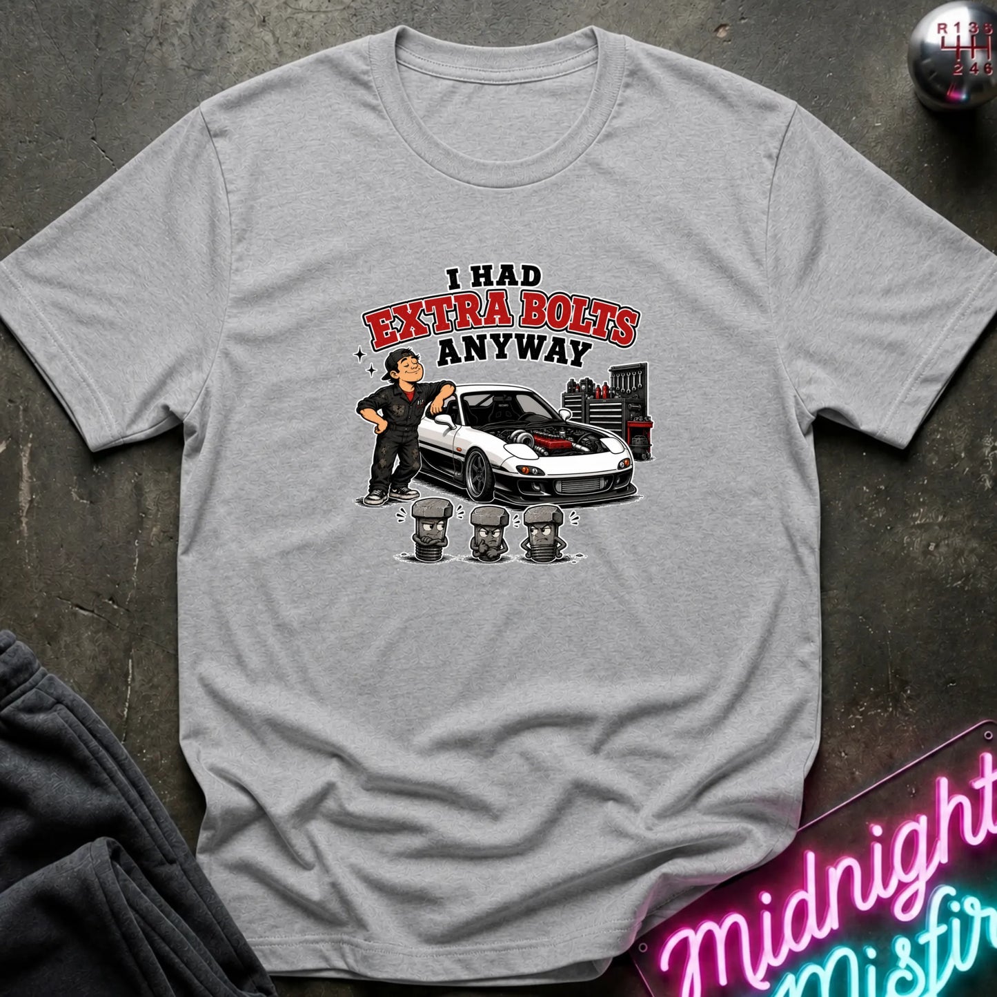 A light gray short-sleeve graphic t-shirt laid flat on a dark textured surface. The graphic features a cartoon of a man standing next to a white sports car with red detailing, with the text 'I HAD EXTRA BOLTS ANYWAY' in red and black.