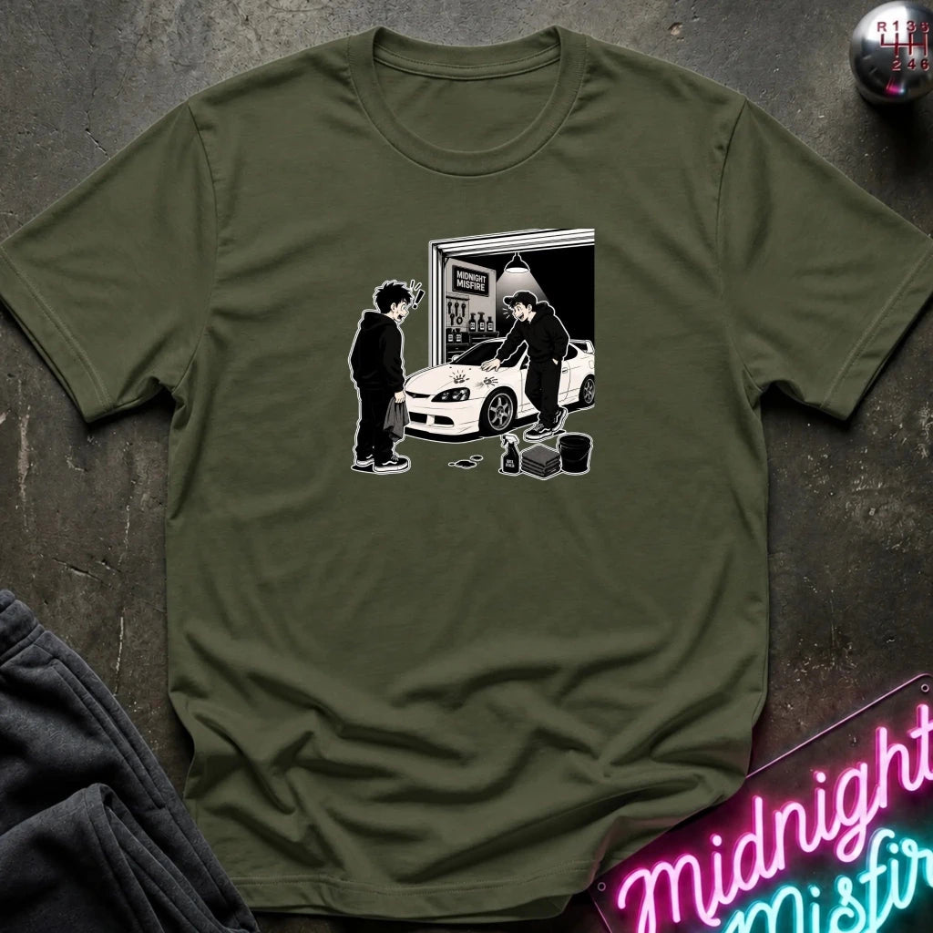 An olive green crewneck t-shirt laid flat on a dark textured surface, featuring a black and white graphic of a person painting a white car in a garage. 