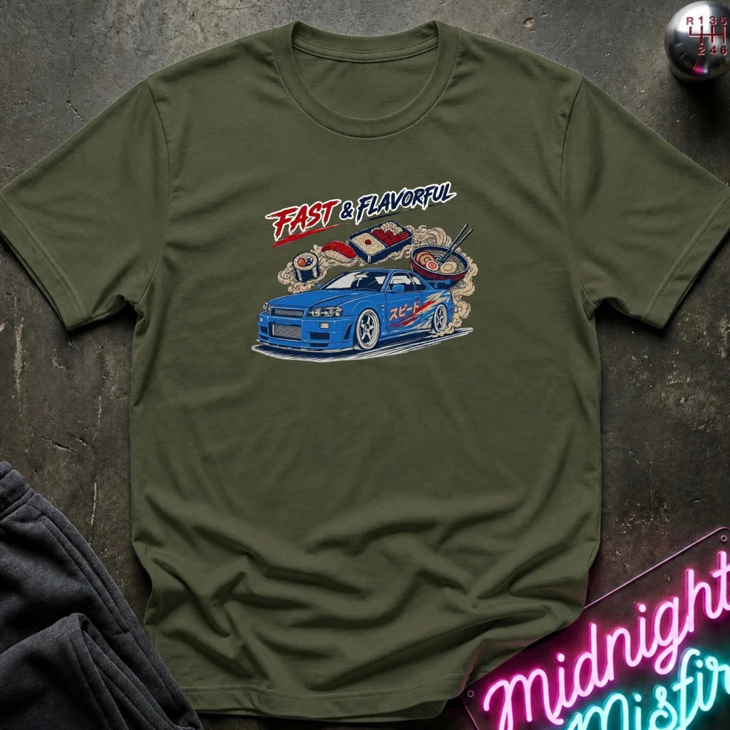 An olive green crewneck t-shirt featuring a graphic print of a blue sports car with smoke and the text 'FAST & FLAVORFUL' in red and white. The shirt is displayed flat on a dark textured surface with a neon sign and dark sweatpants partially visible.