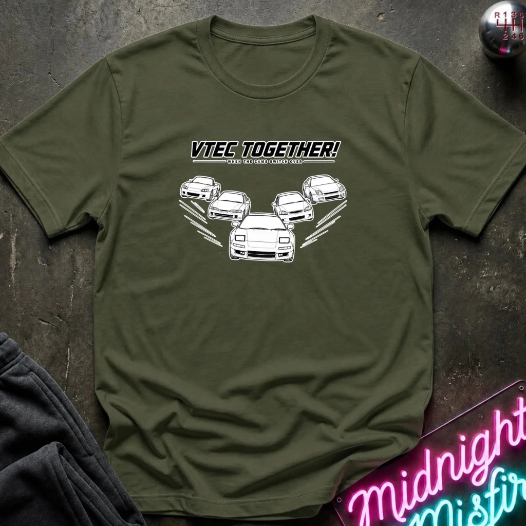 An olive green crewneck t-shirt with a white graphic print of five cars and the text 'VITEC TOGETHER!'. The shirt is laid flat on a dark textured surface with a neon sign and dark pants partially visible in the corners.
