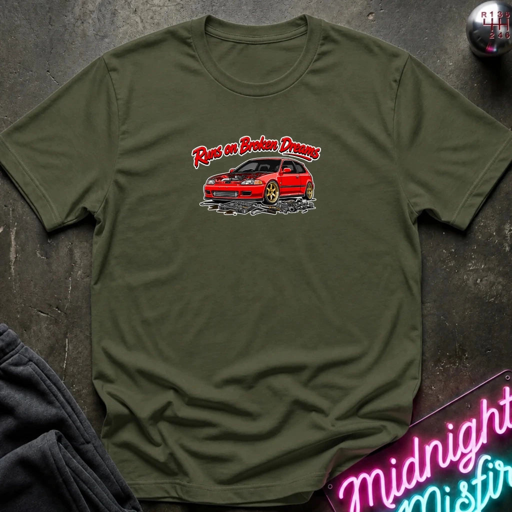 An olive green crewneck t-shirt laid flat on a dark textured surface. The shirt features a graphic print of a red car with the text 'Run on Broken Dreams' in red and white script.
