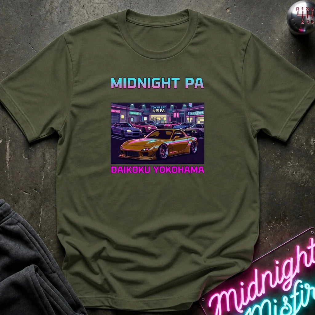 An olive green graphic t-shirt laid flat on a dark textured surface. The shirt features a retro-style neon graphic of a yellow sports car in a parking lot at night, with the text 'MIDNIGHT PA' in a pink-to-blue gradient above and 'DAIKOKU YOKOHAMA' in pink below.