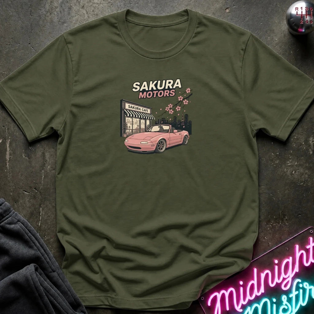 An olive green short-sleeve t-shirt with a graphic print of a pink convertible car in front of a 'Sakura Motors' shop, styled flat on a dark textured surface with a neon sign and dark pants nearby.