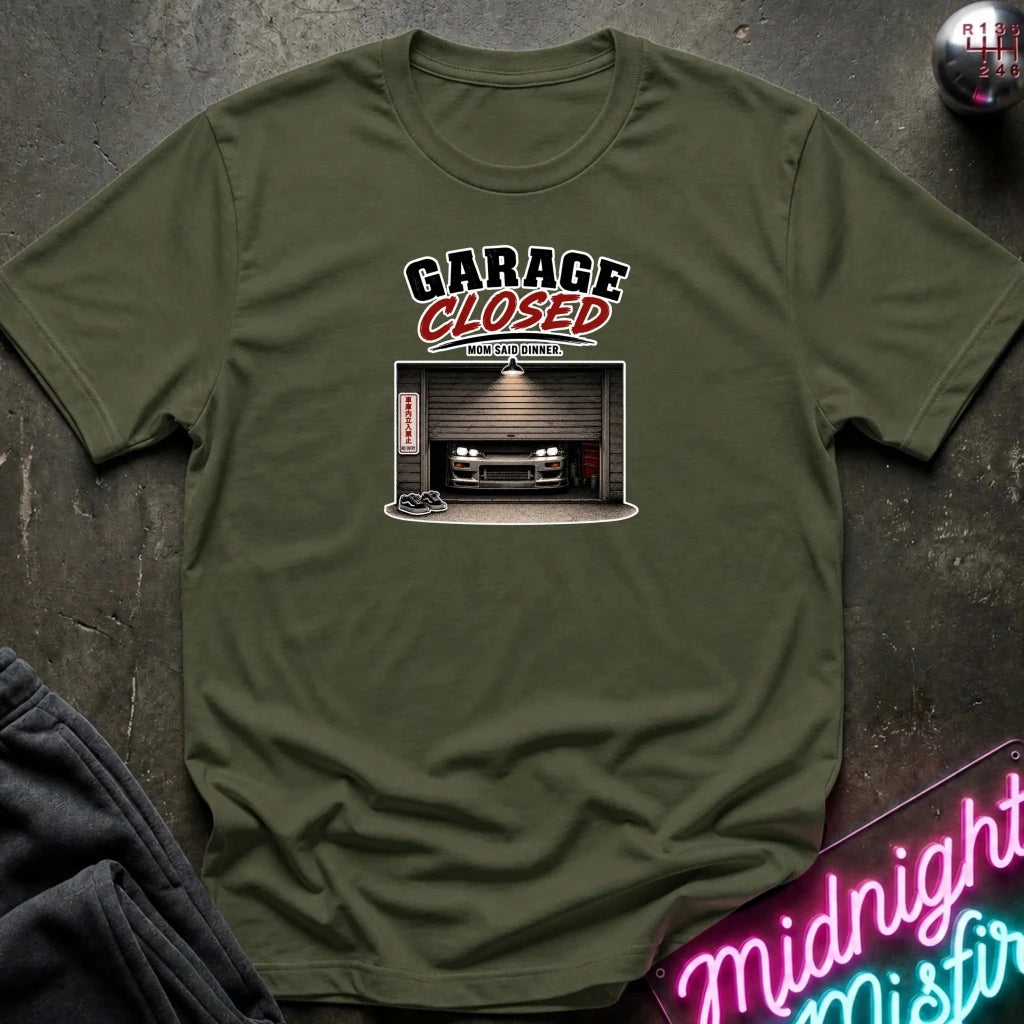 An olive green crewneck t-shirt laid flat on a dark textured surface. The shirt features a graphic print with the text 'GARAGE CLOSED' in black and red, above an illustration of a garage with a car inside.