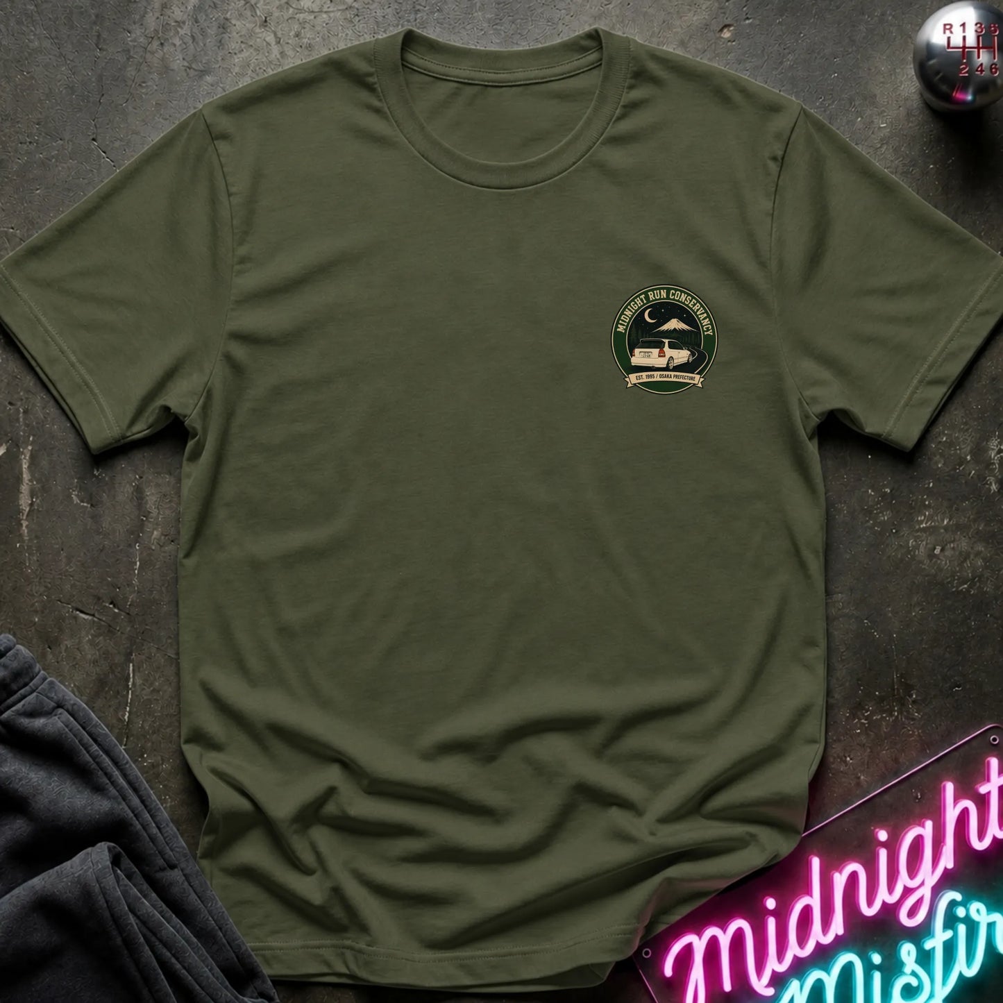 An olive green short-sleeve crewneck t-shirt with a circular green and black chest graphic, styled flat-lay on a dark textured surface with a neon sign and dark pants nearby.