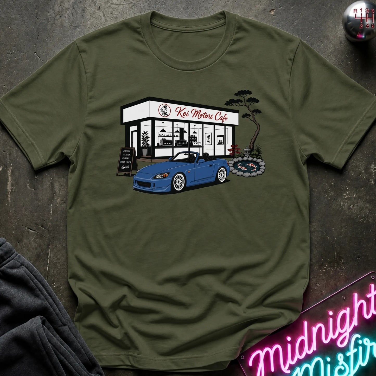 An olive green short-sleeve t-shirt laid flat on a dark textured surface, featuring a graphic print of a blue convertible sports car parked in front of 'Koi Motor Cafe'.