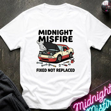 A white short-sleeve graphic T-shirt laid flat on a dark textured surface. The shirt features a printed design of a beige and red car with its hood open, surrounded by wrenches and smoke, with the text 'MIDNIGHT MISFIRE' above and 'FIXED NOT REPLACED' below.