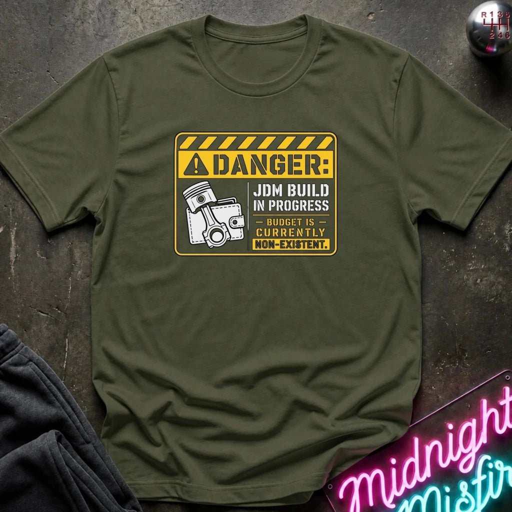 An olive green crewneck t-shirt laid flat on a dark textured surface, featuring a yellow and black warning sign graphic with a jackhammer and the text 'DANGER: JDM BUILD IN PROGRESS - BUDGET IS CURRENTLY NON-EXISTENT.'.