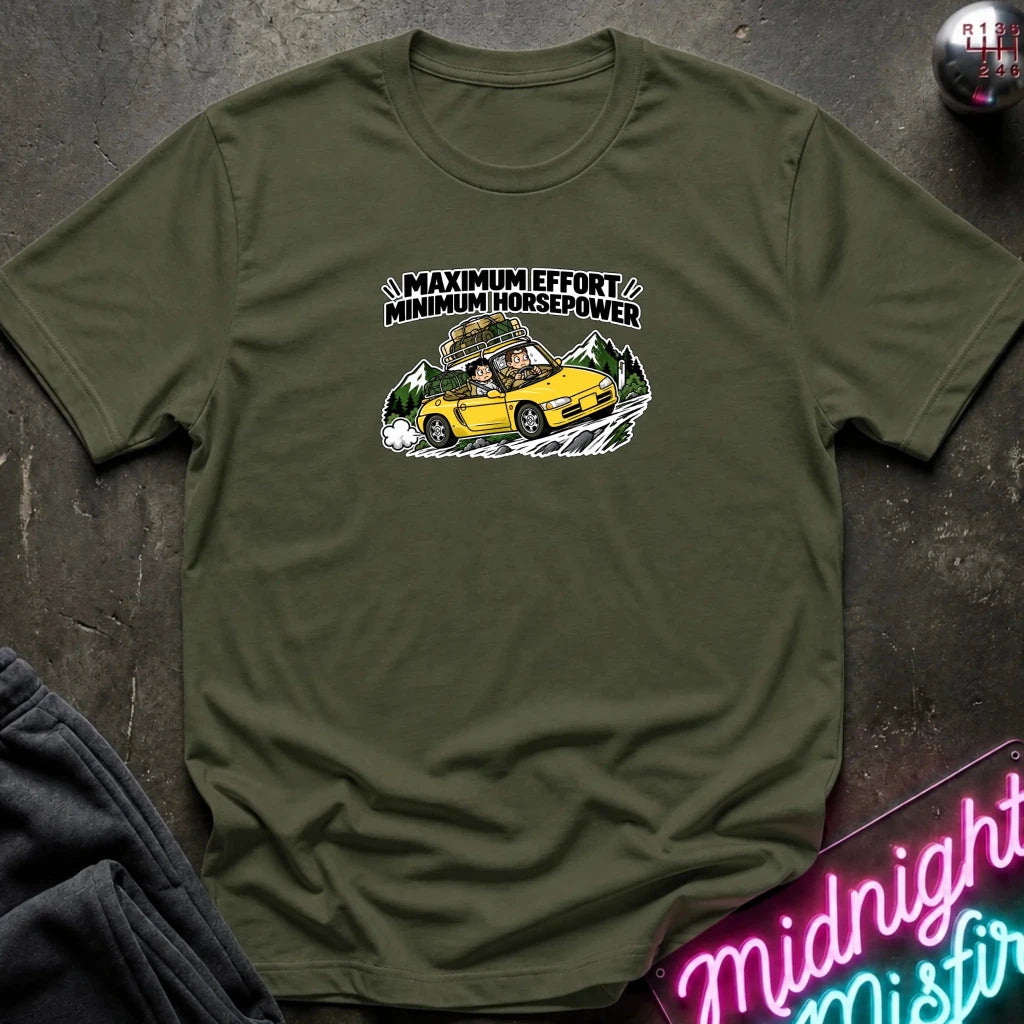 olive graphic t-shirt laid flat on a dark textured surface. The graphic features a yellow sports car with a driver and passengers, with the text 'MAXIMUM EFFORT MINIMUM HORSEPOWER' above it.