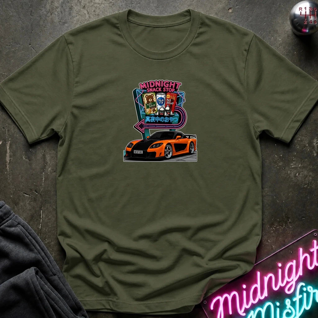 An olive green  short-sleeve t-shirt laid flat on a dark textured surface, featuring a colorful graphic print of an orange sports car in front of a neon sign that reads 'Midnight Snack Stop'.