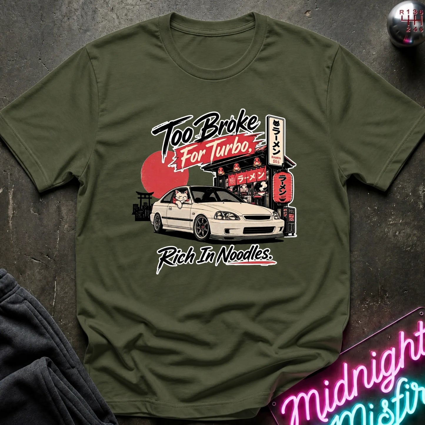 Olive green short-sleeve graphic T-shirt laid flat on a dark textured surface. The shirt features a printed design of a white sports car in front of a Japanese-style street scene with a red sun and neon signs, plus the text 'Too Broke For Turbo' and 'Rich In Noodles.